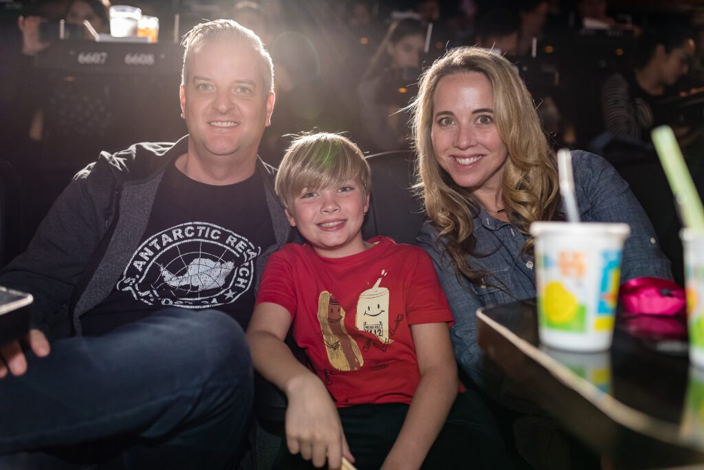 A man and woman sit together with a child in a movie theater, enjoying a film in a dimly lit environment.